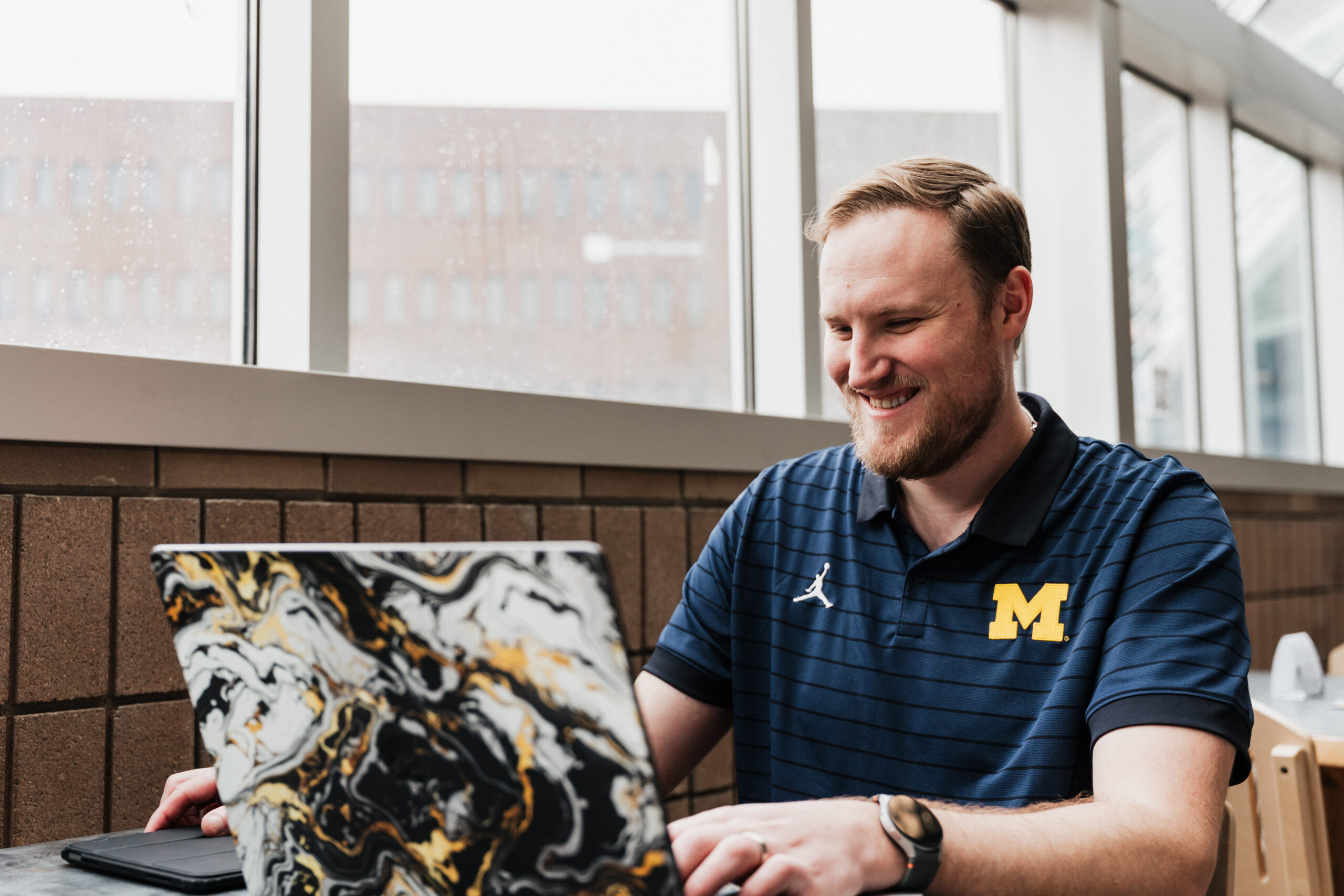 A person wearing a blue University of Michigan polo shirt working on a laptop with a black, white, and gold marbled case, sitting by a window.