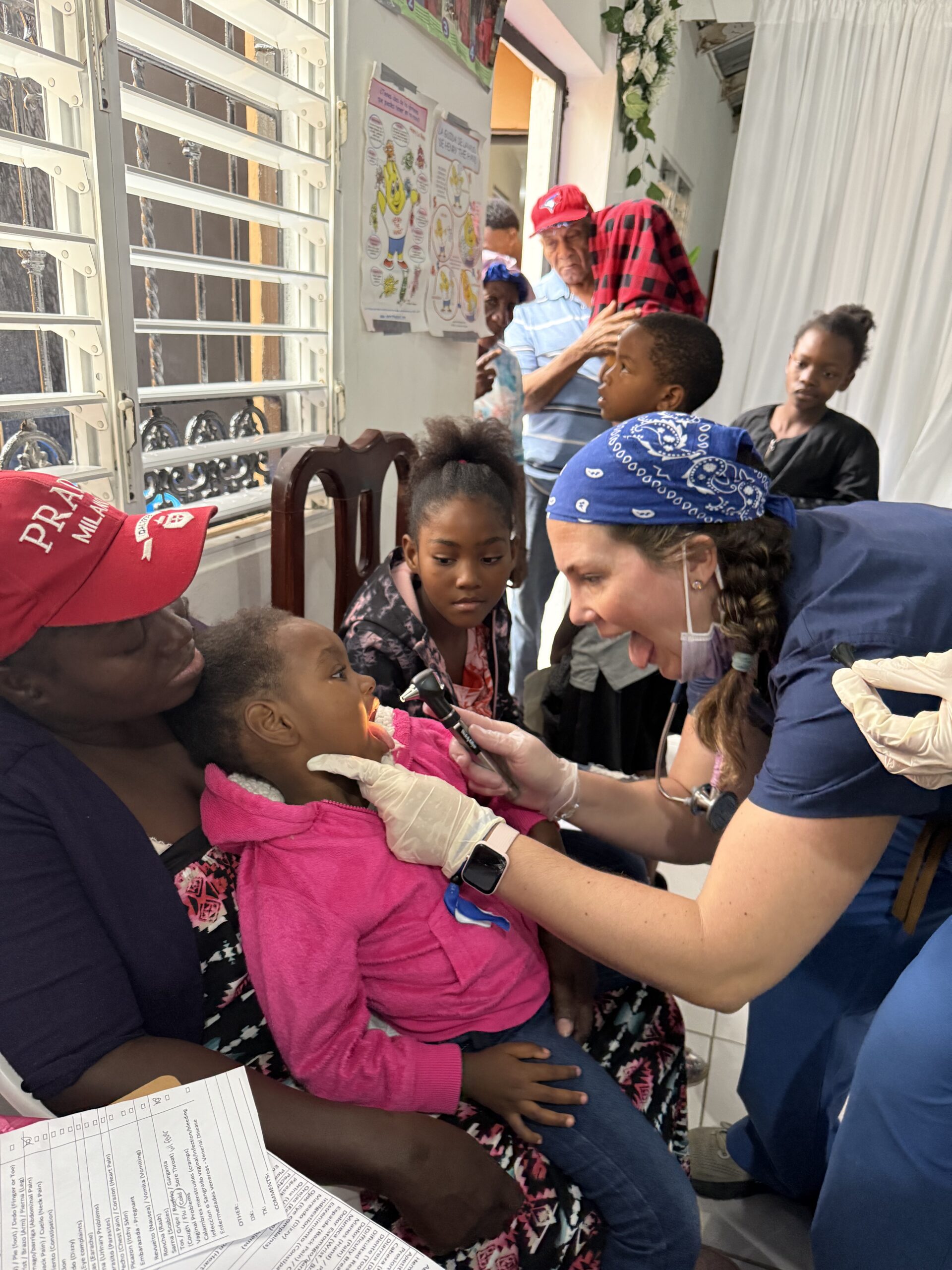 A health care worker wearing gloves and a blue bandana uses an otoscope to examine a young child sitting on an adult's lap in a clinic with other people waiting.