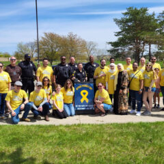 Group of people, including police officers and civilians wearing yellow "Officer Friendly Day" t-shirts, posing outdoors with a sign that reads "Officer Friendly Day. Do you have any safety questions? Ask me!"