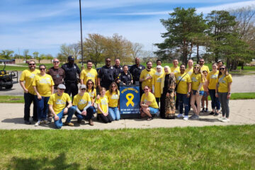 Group of people, including police officers and civilians wearing yellow "Officer Friendly Day" t-shirts, posing outdoors with a sign that reads "Officer Friendly Day. Do you have any safety questions? Ask me!"