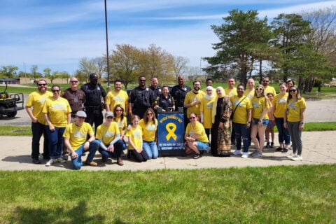 Group of people, including police officers and civilians wearing yellow "Officer Friendly Day" t-shirts, posing outdoors with a sign that reads "Officer Friendly Day. Do you have any safety questions? Ask me!"