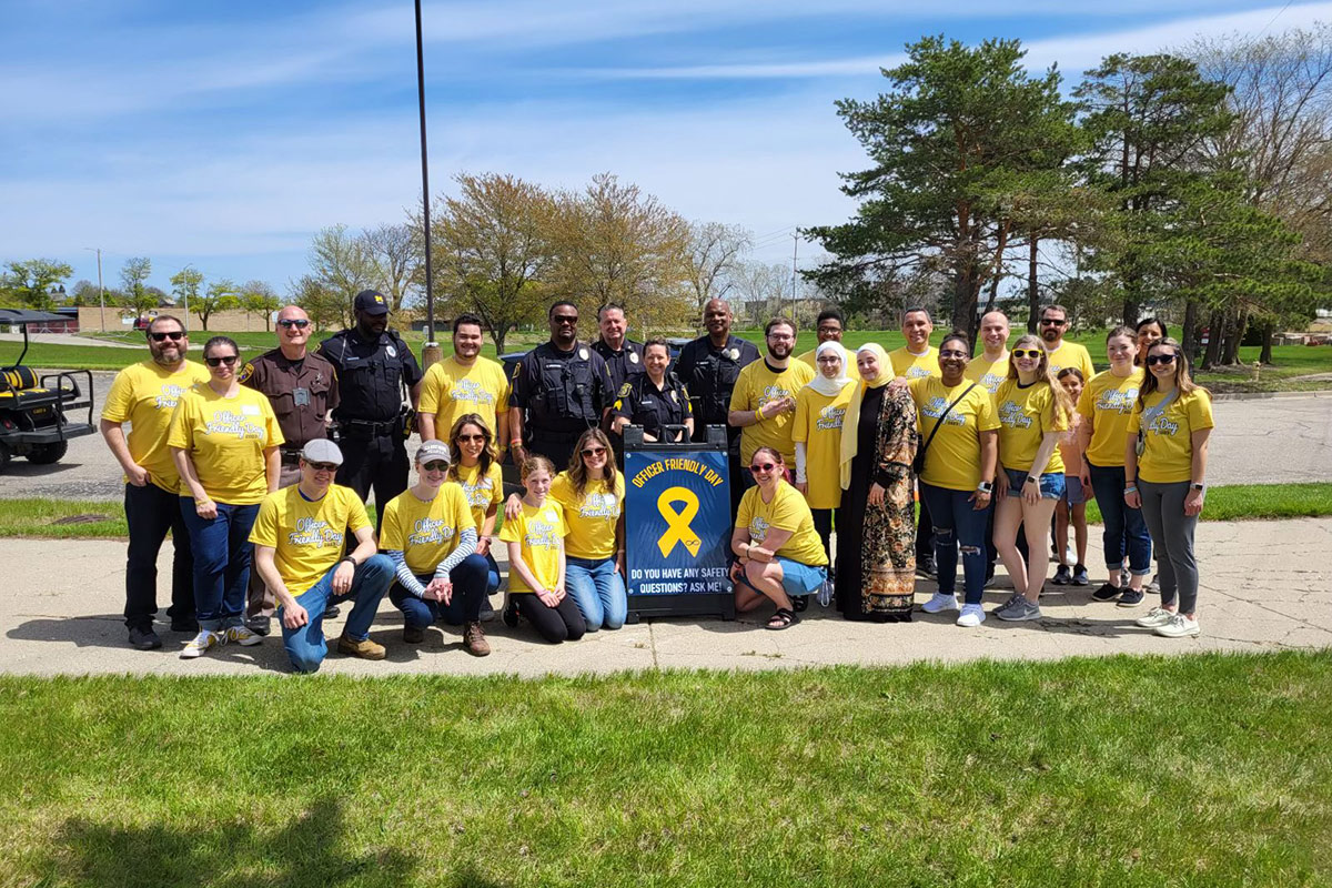 Group of people, including police officers and civilians wearing yellow "Officer Friendly Day" t-shirts, posing outdoors with a sign that reads "Officer Friendly Day. Do you have any safety questions? Ask me!"