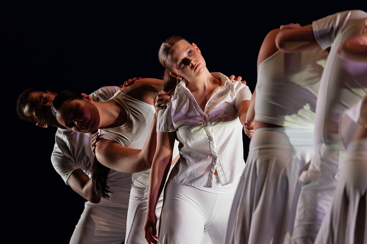 A group of dancers dressed in white performing a contemporary dance routine against a dark background.