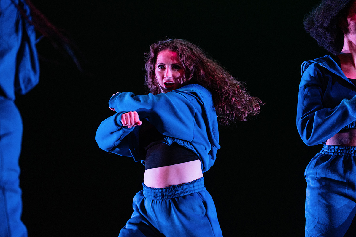 Three dancers in blue outfits performing against a black background, with one dancer's curly hair in motion.