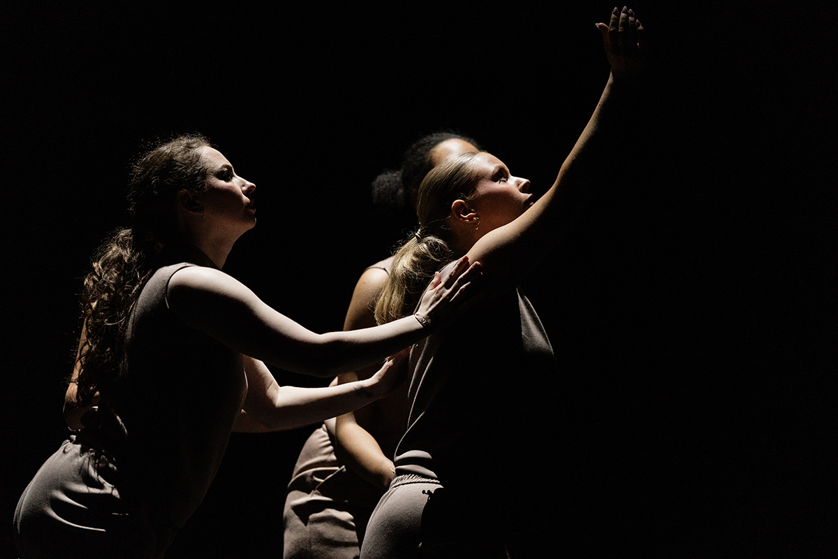 Three dancers in dim light, one reaching upward while another supports her back.