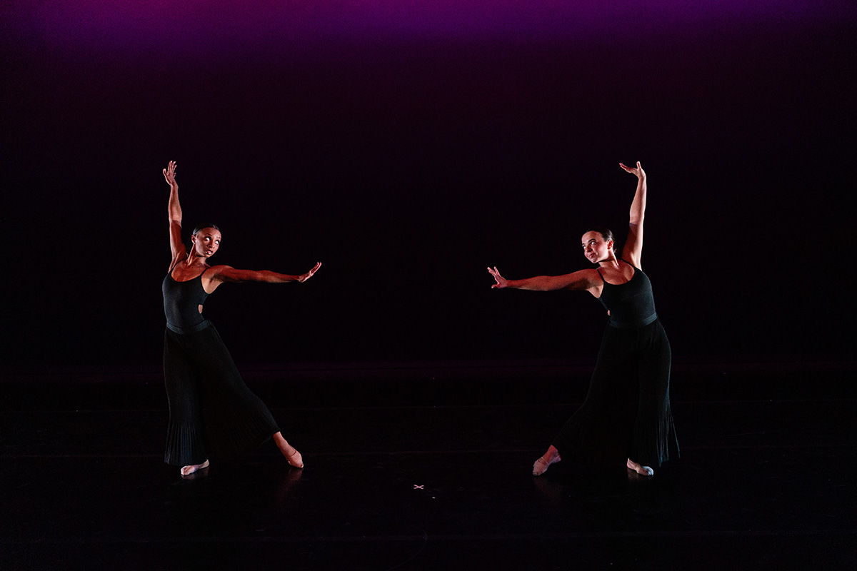 Two dancers in black outfits performing a contemporary dance on a dark stage with purple lighting.