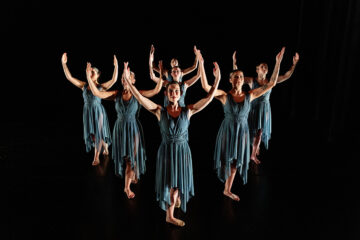 Seven dancers in blue dresses perform a synchronized pose with arms raised on a dark stage.