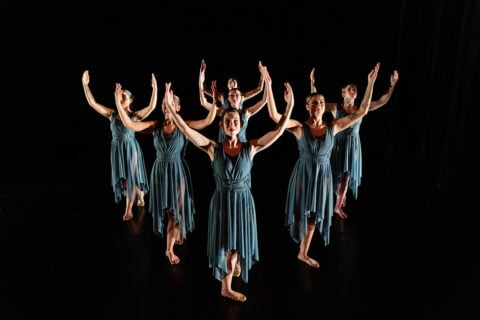 Seven dancers in blue dresses perform a synchronized pose with arms raised on a dark stage.