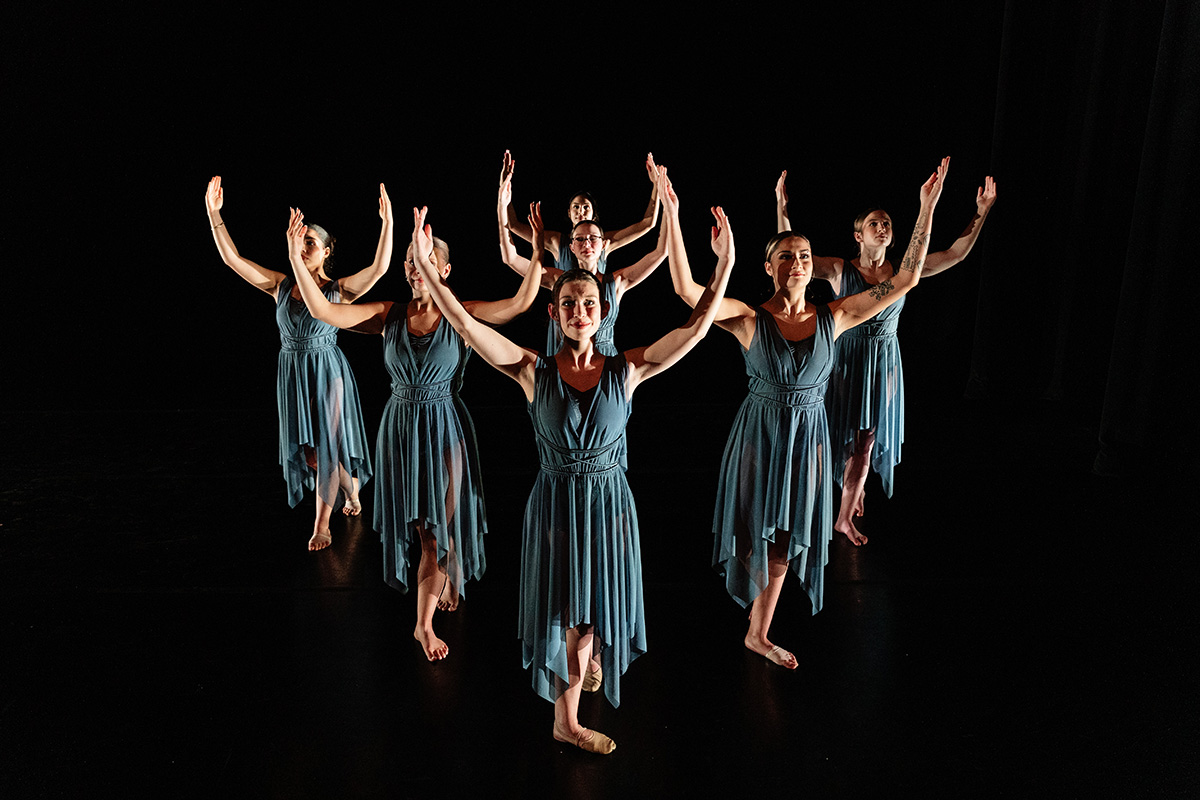 Seven dancers in blue dresses perform a synchronized pose with arms raised on a dark stage.