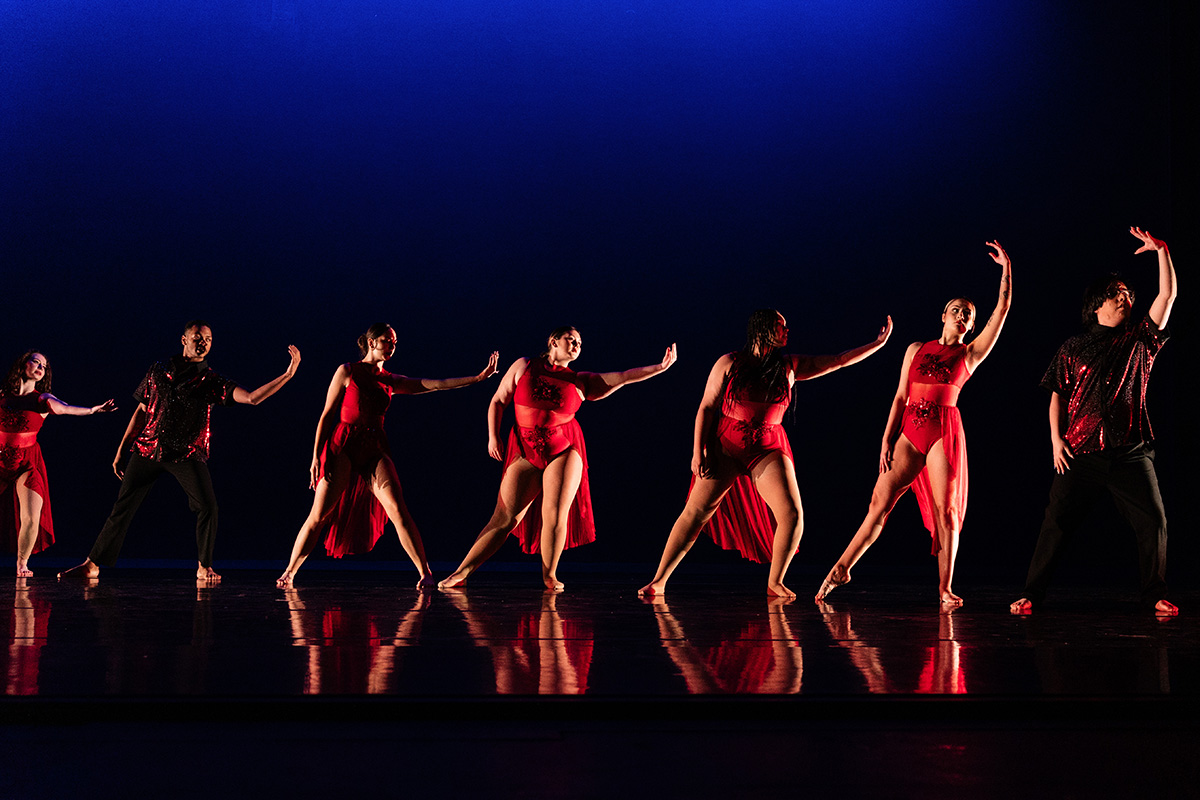 Seven dancers in red costumes perform a synchronized dance on a dark stage with blue lighting.