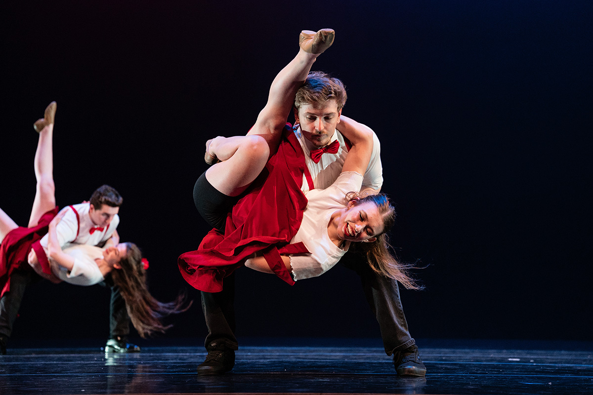 Two couples performing an acrobatic dance lift on stage, wearing red and white costumes against a dark background.