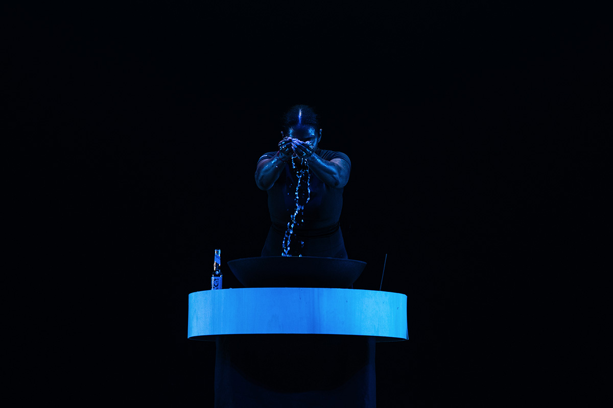 Person standing behind a round table with a large bowl, pouring water from their hands into the bowl in a dark setting with blue lighting.