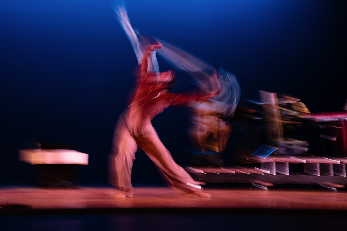 Blurred image of a dancer in motion on stage with a dark blue background and musical instruments in the background.