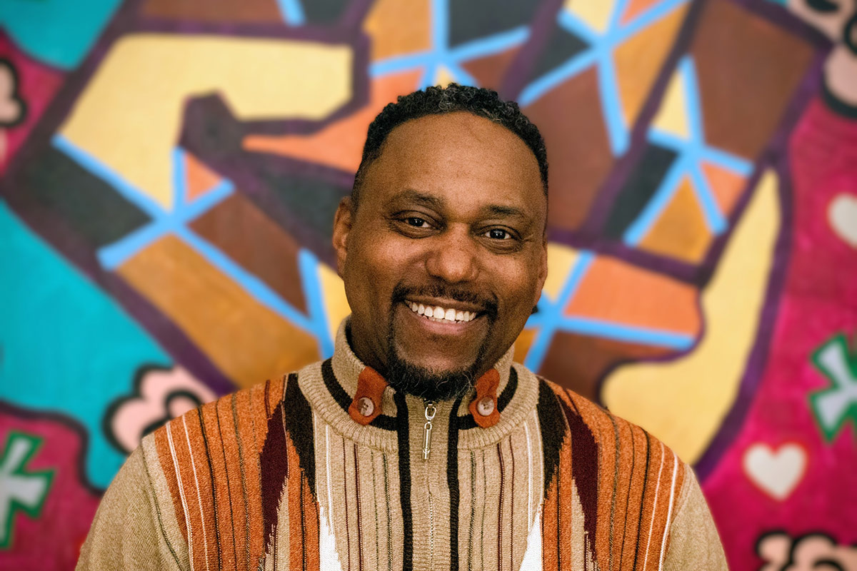 Person wearing a multicolored striped sweater standing in front of a colorful graffiti wall with geometric shapes and hearts.