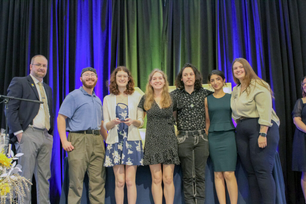 Group of seven people standing on a stage with blue and green curtains, one person holding an award.