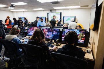 People playing video games on computers in a busy gaming room with multiple monitors and others standing in the background.