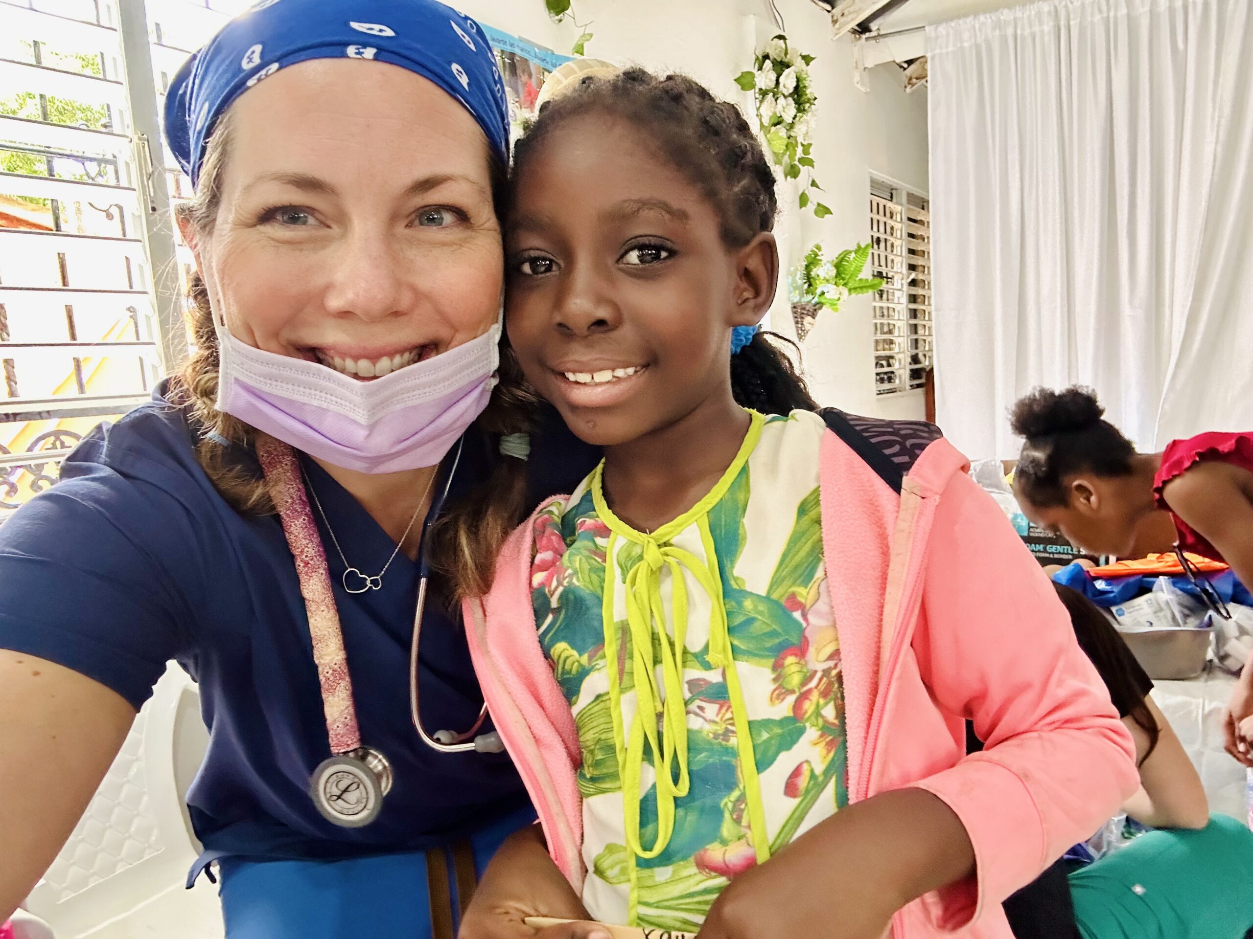 A healthcare worker wearing navy scrubs and a blue bandana with a stethoscope around their neck sits next to a child in a pink jacket and floral shirt inside a bright room.