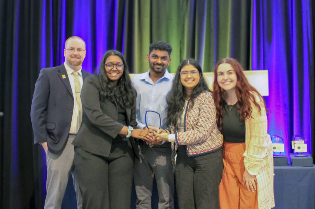 Five people standing together on a stage holding an award, with a backdrop of blue and green curtains.