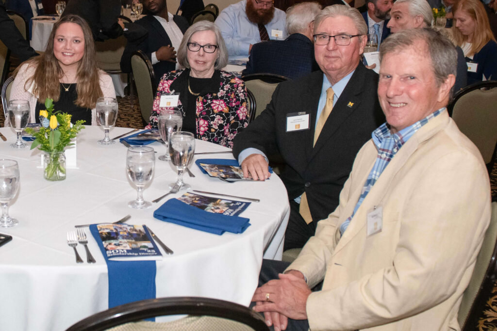 Four people sitting at a round banquet table set with a white tablecloth, water glasses, blue napkins, and event programs.