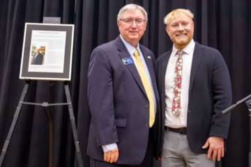 Two men in suits standing side by side in front of a black curtain with a framed document on an easel to the left.