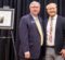 Two men in suits standing side by side in front of a black curtain with a framed document on an easel to the left.