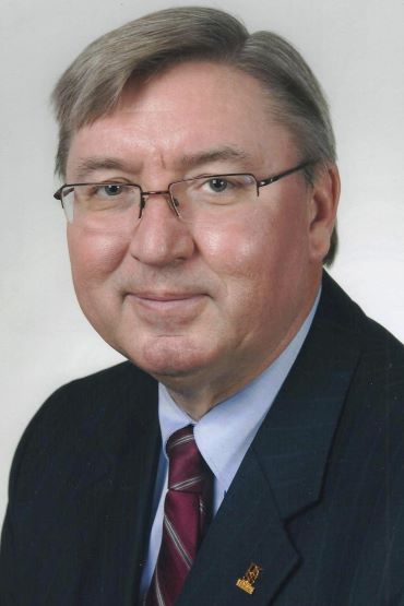 A portrait of a man wearing glasses, a dark suit, light-blue shirt, and red striped tie against a plain background.