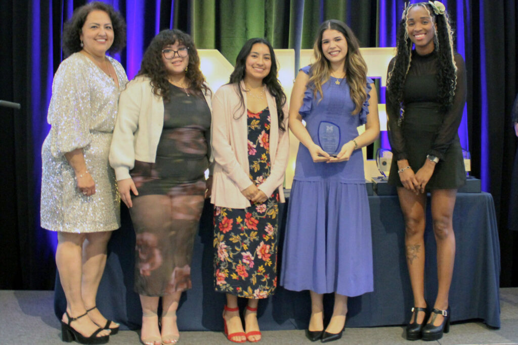 Five women standing in a row at an event, one holding an award plaque.