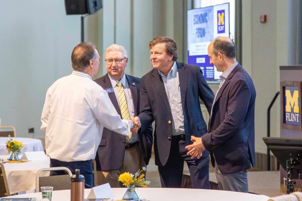 Four men in business attire shaking hands and conversing at a University of Michigan-Flint event.