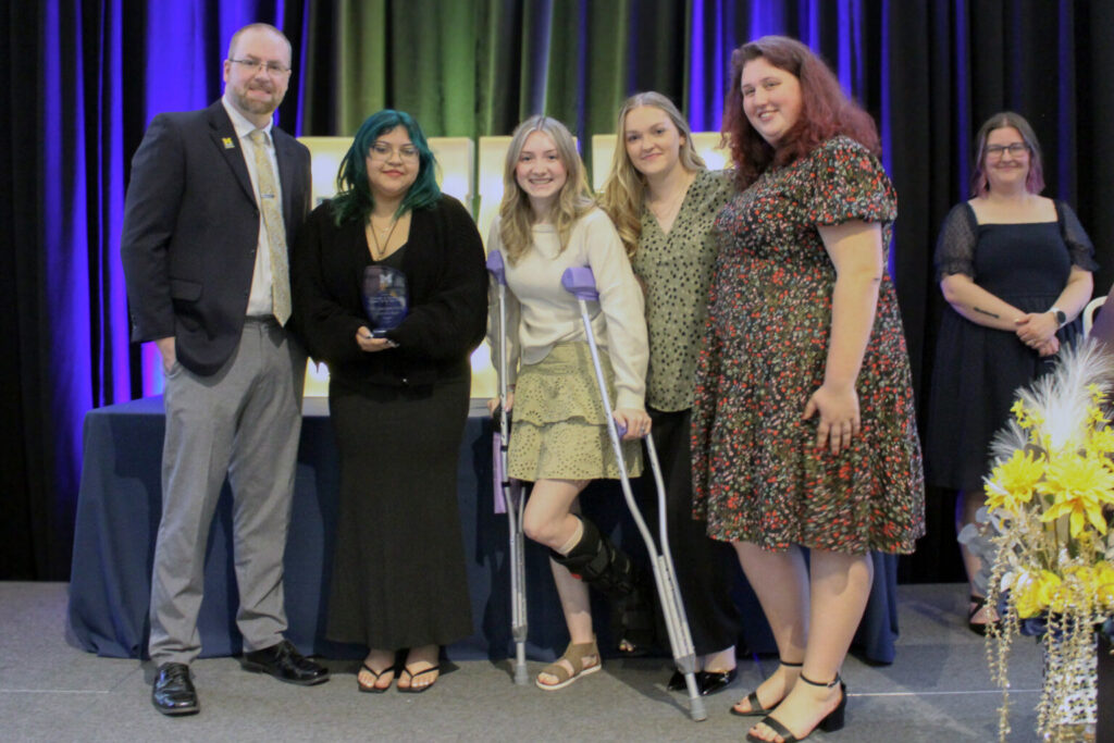 Group of six people standing on a stage with blue and green curtains, one person holding an award, one person using crutches and wearing a leg brace.