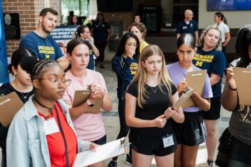 Group of people standing indoors, some holding clipboards and wearing name tags, with a few wearing Michigan Anesthesia shirts.