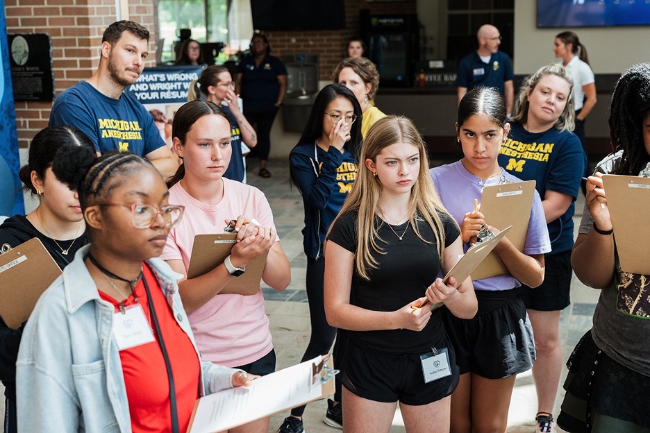 Group of people standing indoors, some holding clipboards and wearing name tags, with a few wearing Michigan Anesthesia shirts.