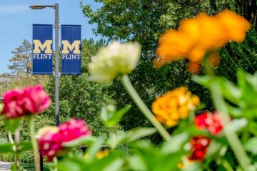 Two blue banners with a yellow "M" and the word "Flint" hang from a streetlamp, with colorful flowers and green trees in the foreground.