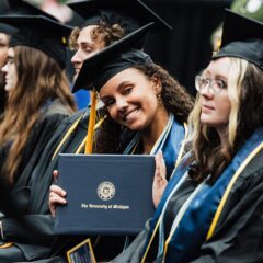 Group of graduates in caps and gowns, one holding a University of Michigan diploma cover.