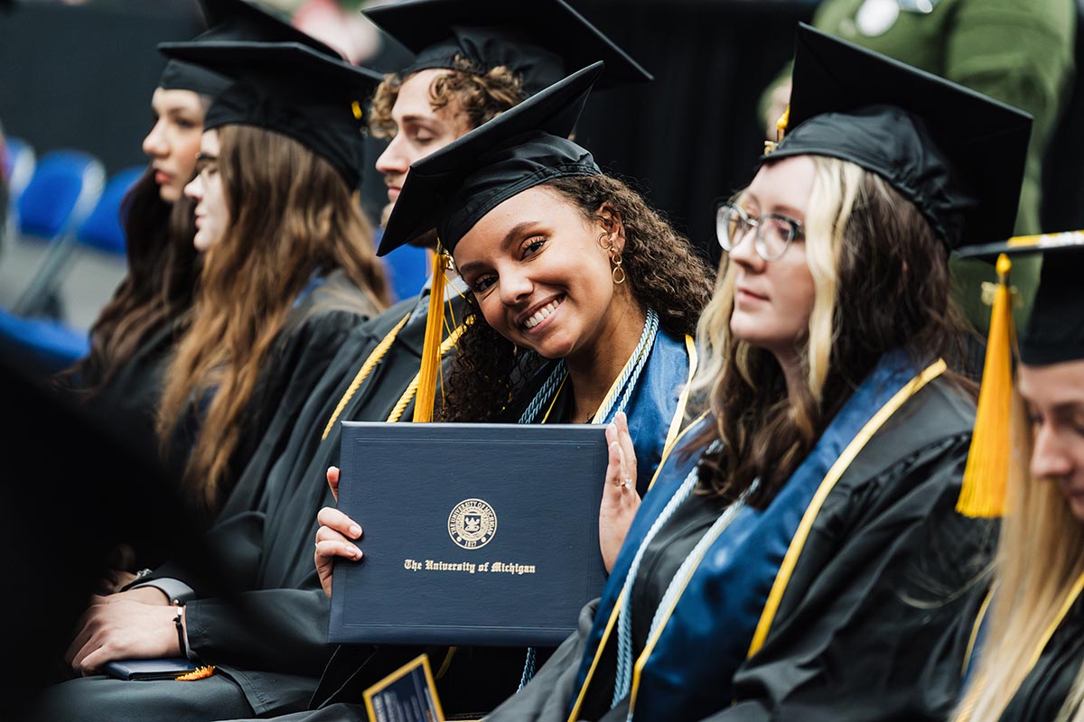 Group of graduates in caps and gowns, one holding a University of Michigan diploma cover.