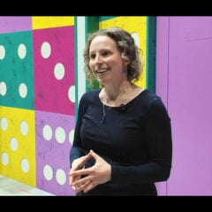 Person standing in front of a colorful wall with large domino patterns, wearing a black long-sleeve shirt and a necklace.