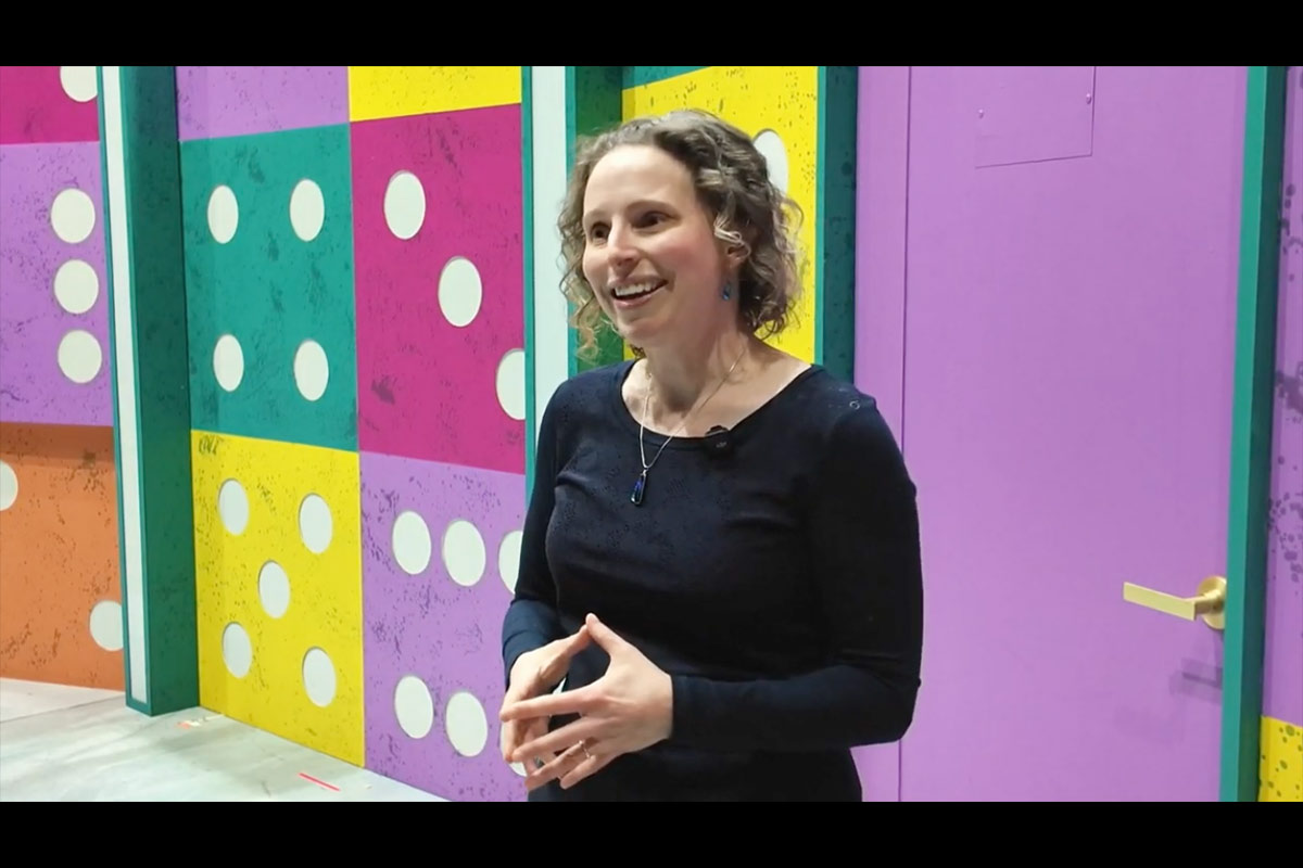 Person standing in front of a colorful wall with large domino patterns, wearing a black long-sleeve shirt and a necklace.