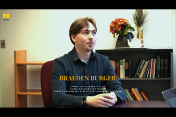 Person sitting at a desk with text overlay reading "Braeden Burger, Computer Science Major, Music Composition Minor, Winter 2026 Maize & Blue Distinguished Scholar," with a bookshelf and flowers in the background and a University of Michigan-Flint logo in the top left corner.