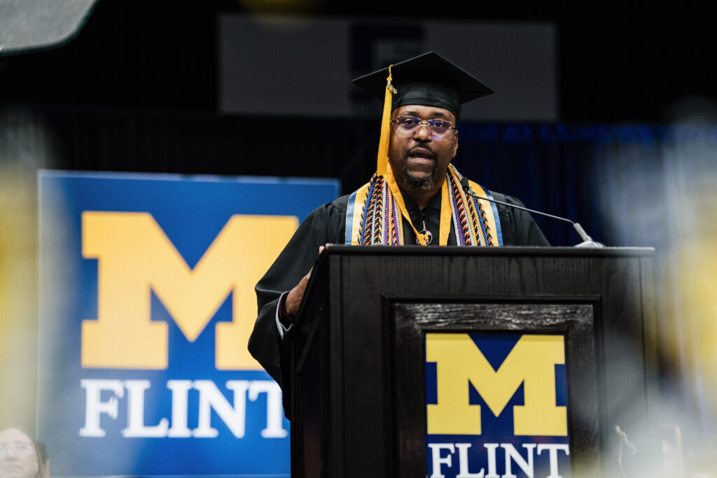 Ken Whittaker speaking at the lectern with the UM-Flint logo featured prominently. 