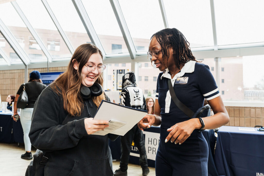Two young women talking indoors at an event with tables and informational materials in the background.