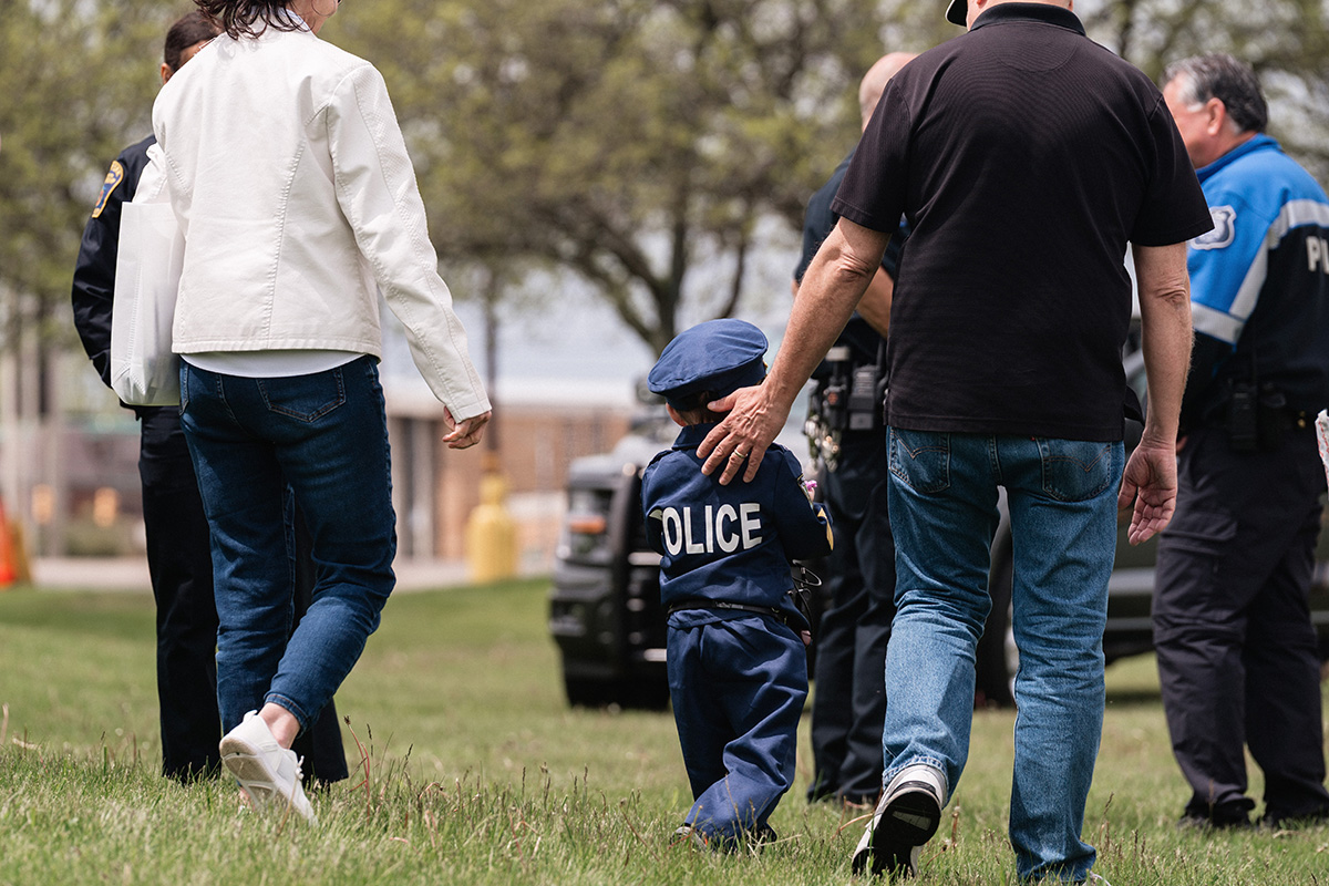 A child dressed in a police uniform walking on grass, accompanied by adults and police officers.