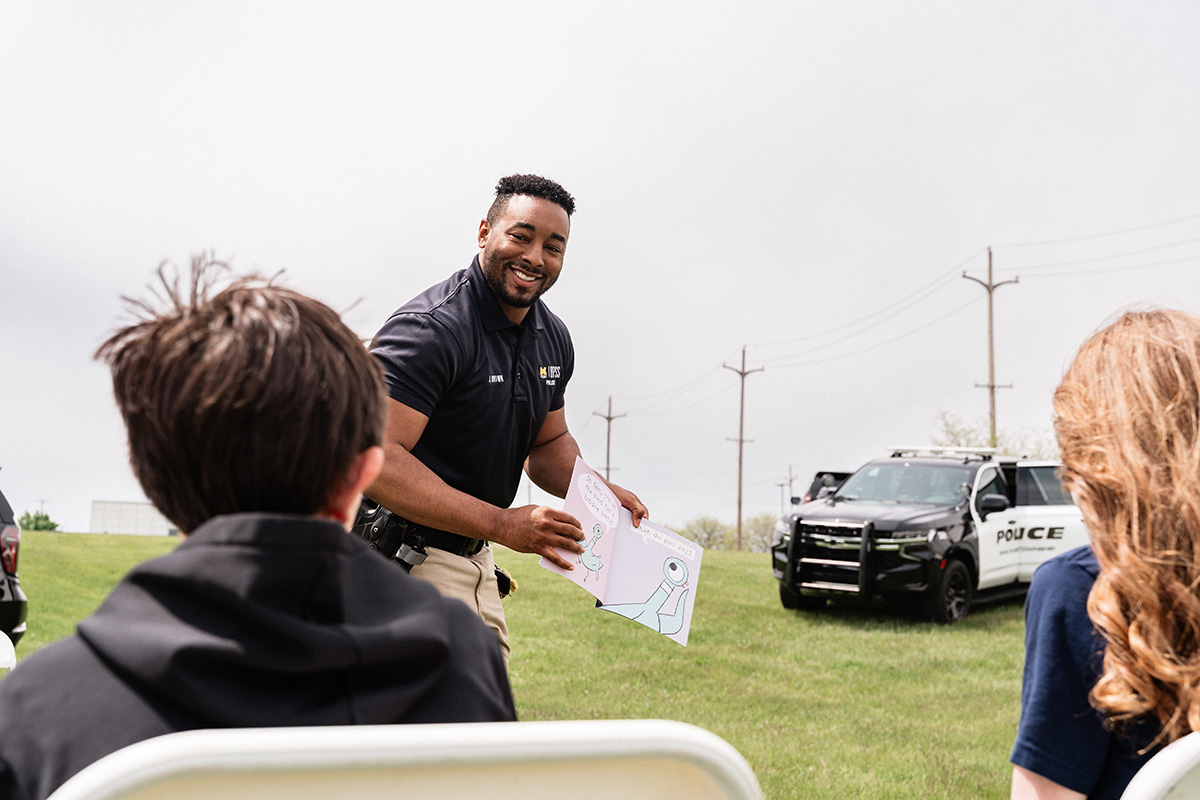 Police officer reading a children's book outdoors to two seated children with a police car in the background.
