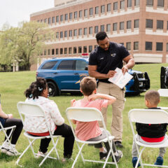 A police officer is standing and showing a booklet to a group of children seated on white folding chairs outdoors on grass, with police vehicles and a building in the background.
