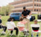 A police officer is standing and showing a booklet to a group of children seated on white folding chairs outdoors on grass, with police vehicles and a building in the background.