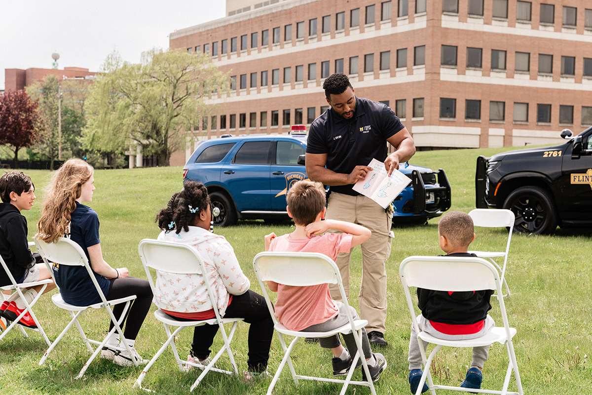 A police officer is standing and showing a booklet to a group of children seated on white folding chairs outdoors on grass, with police vehicles and a building in the background.