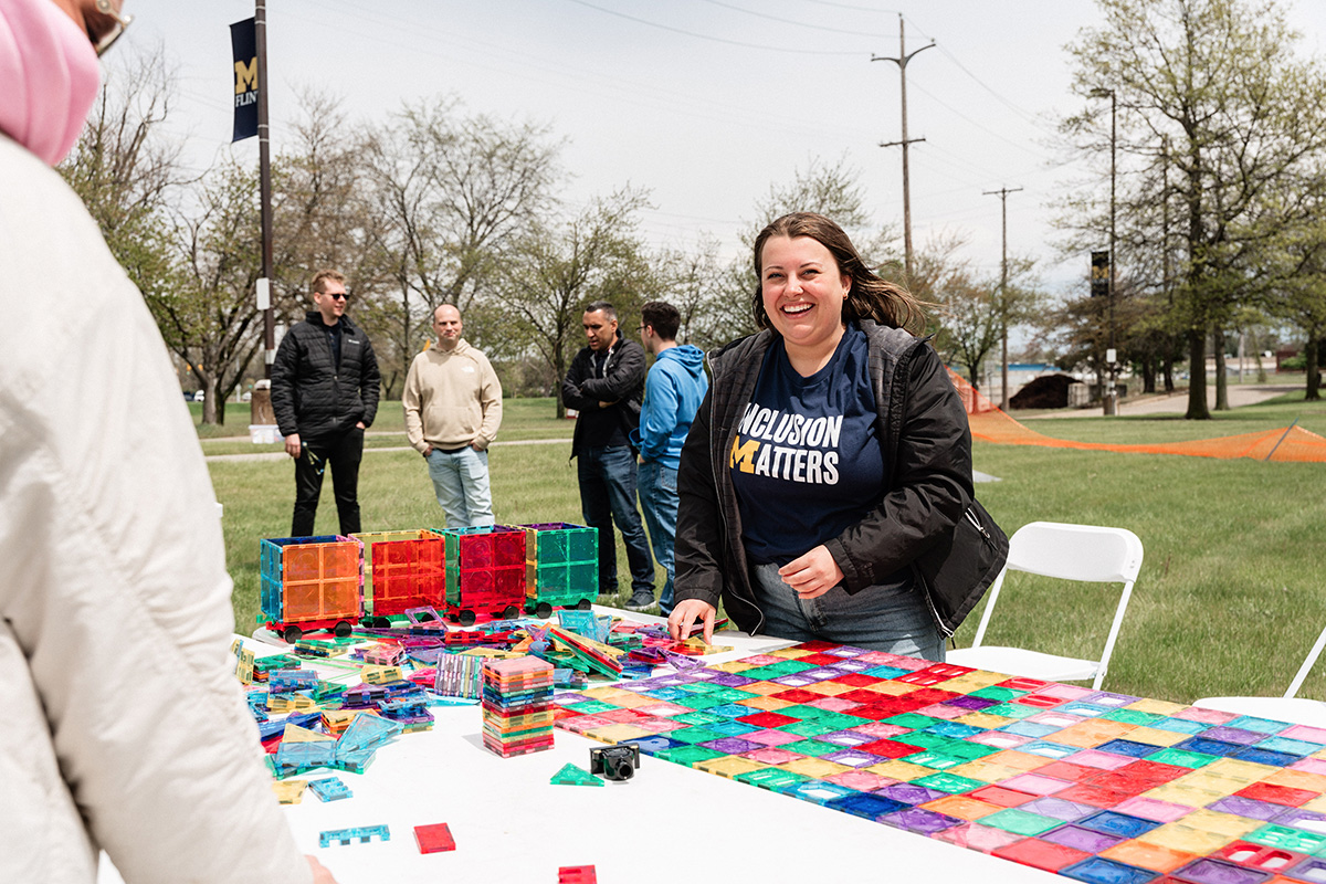 Person wearing a navy shirt with "Inclusion Matters" arranging colorful translucent tiles on a table outdoors with others in the background.