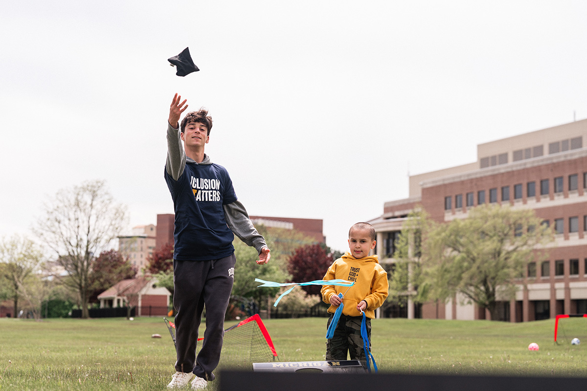 Two people playing cornhole outdoors on a grassy field with buildings and trees in the background.