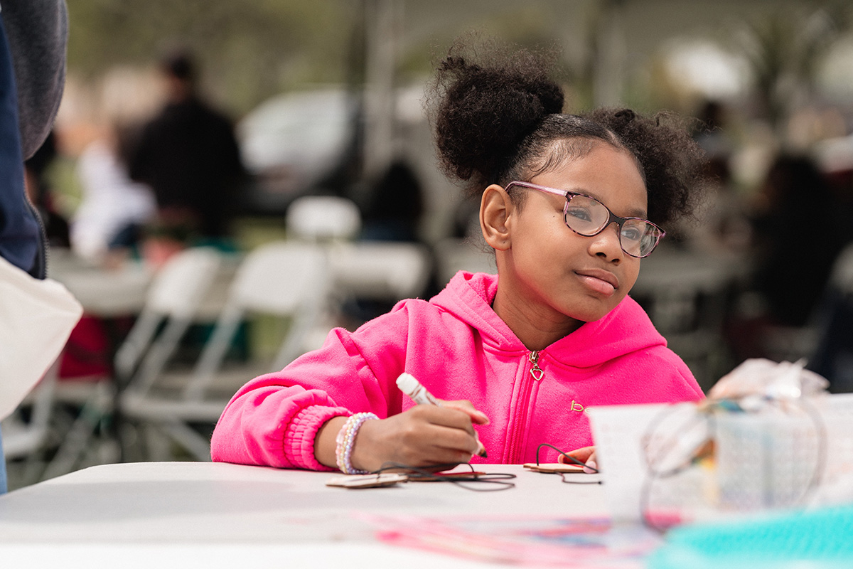 Child in a bright pink hoodie writing or drawing at a table outdoors with blurred people and chairs in the background.