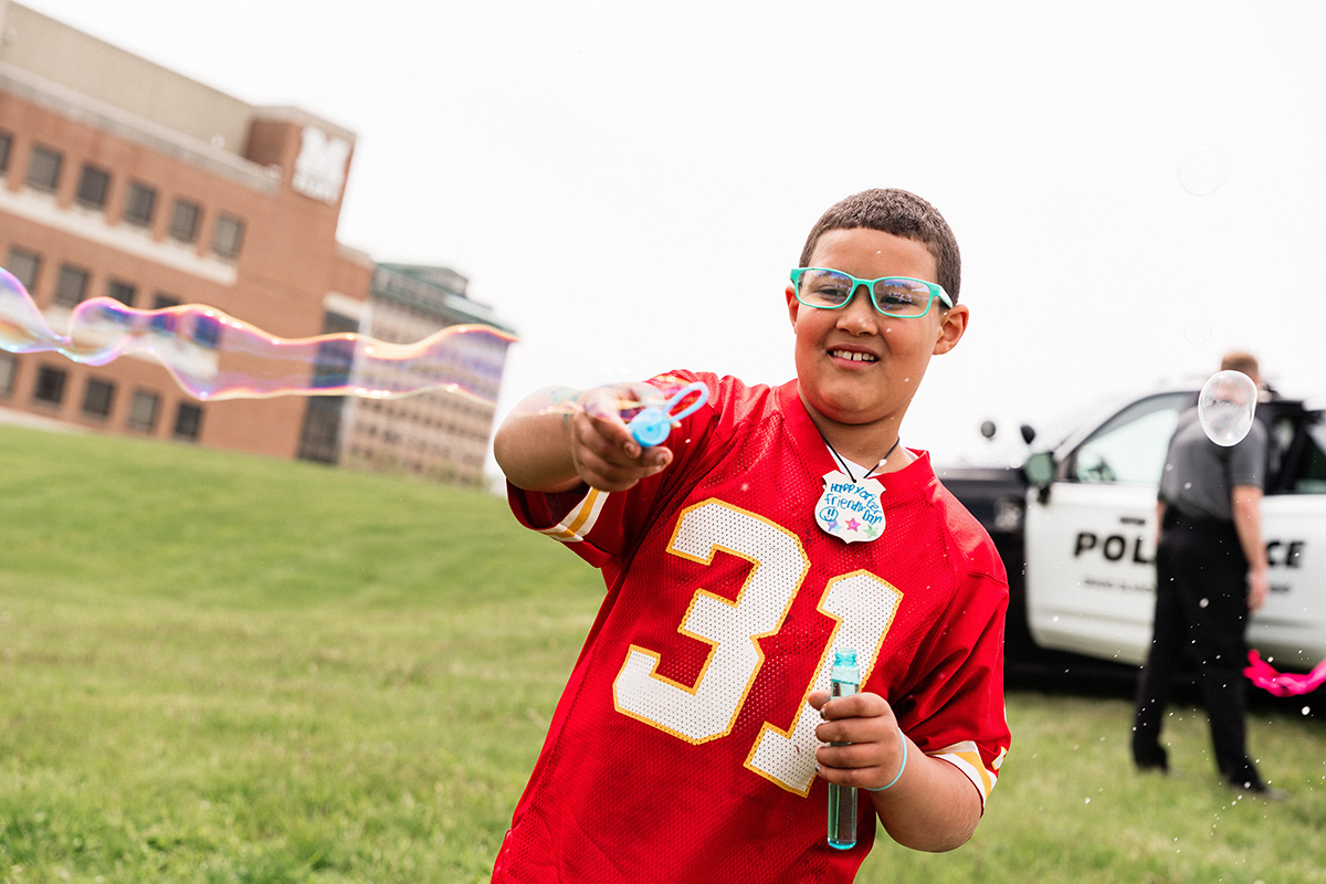 Child in a red sports jersey blowing bubbles outdoors near a police car and officer.