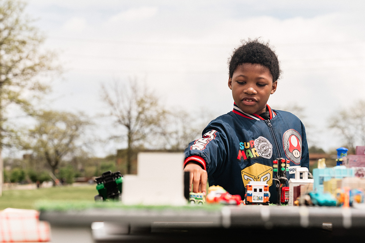 Child playing with toy cars and buildings on a table outdoors.