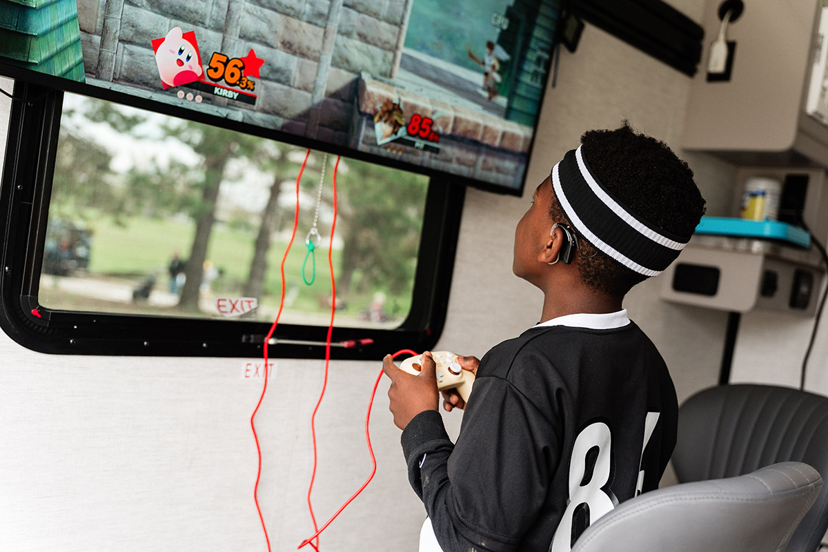 A boy wearing a black jersey and headband plays a video game on a wall-mounted TV inside a room.